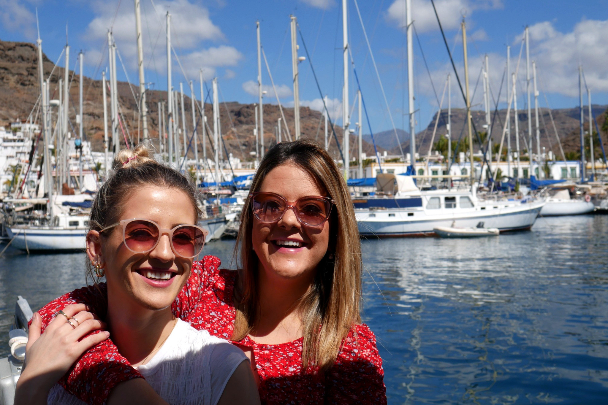 Two friends smiling on a boat in Puerto de Mogan marina with yachts and mountains in the background, Gran Canaria